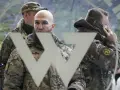 Trzech mężczyzn w mundurach, jeden rozmawia przez telefon, na tle symbolu przypominającego literę "W". Mogą to być członkowie grupy Wagnera.