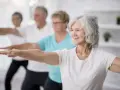 Seniorzy ćwiczą, by zapobiegać sarkopenii. Uśmiechnięta kobieta na pierwszym planie wykonuje ćwiczenie z wyciągniętymi rękami.