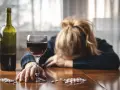 Kobieta z głową opartą na rękach, obok kieliszek czerwonego wina, butelka i opakowania leków, w tym Clexane. Alkohol i leki na stole.