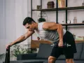 Mężczyzna wykonuje trening siłowy w domu, podnosząc hantle. Opiera się o krzesło, skupiony na ćwiczeniu.