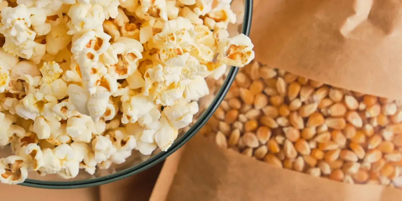 Misternie przygotowany popcorn w szklanej misce obok ziaren kukurydzy w papierowej torbie. Czy popcorn tuczy? Zależy od dodatków!