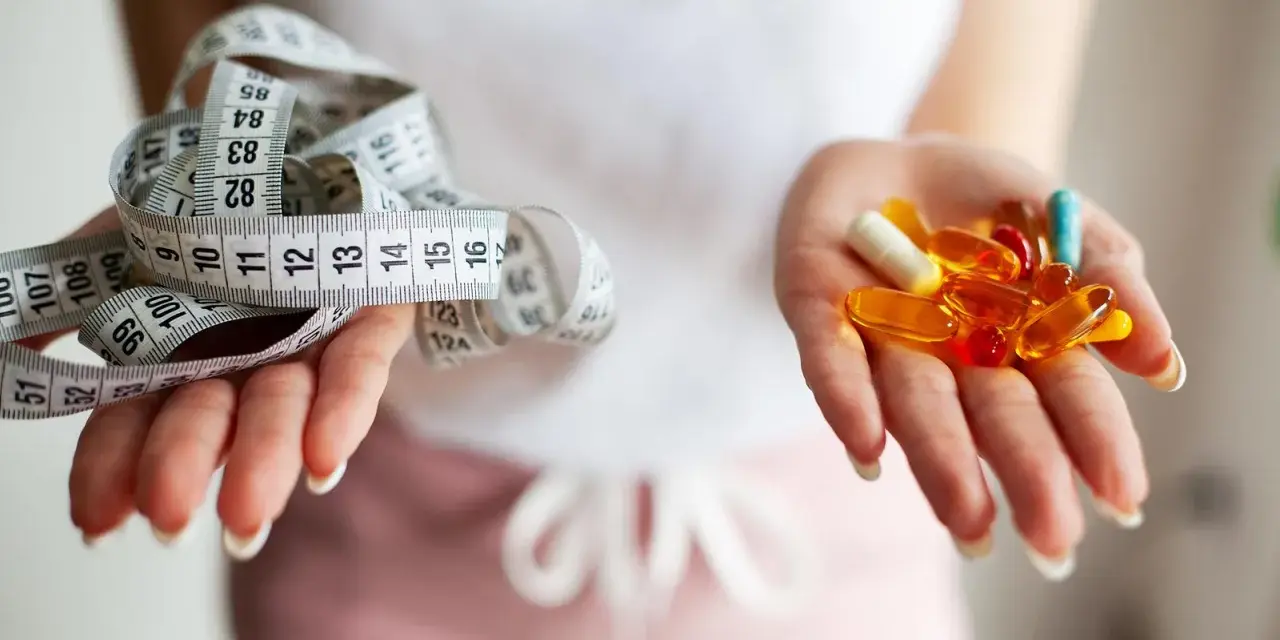 Dwie dłonie: jedna z miarką krawiecką, druga z kolorowymi kapsułkami – symbolizują wybór między dietą a środkami na odchudzanie.