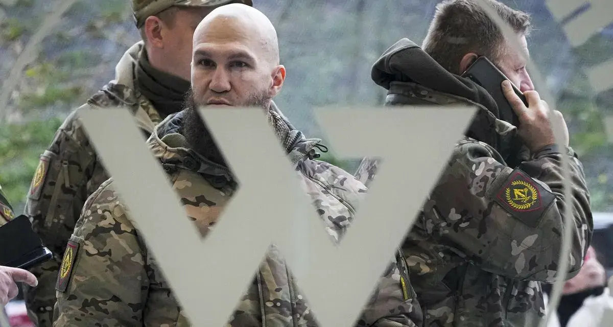 Trzech mężczyzn w mundurach, jeden rozmawia przez telefon, na tle symbolu przypominającego literę "W". Mogą to być członkowie grupy Wagnera.