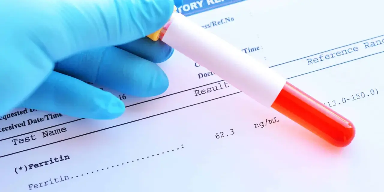 Ręka w niebieskiej rękawiczce trzyma probówkę z krwią nad raportem laboratoryjnym. Wynik badania ferrytyny to 62.3 ng/mL.