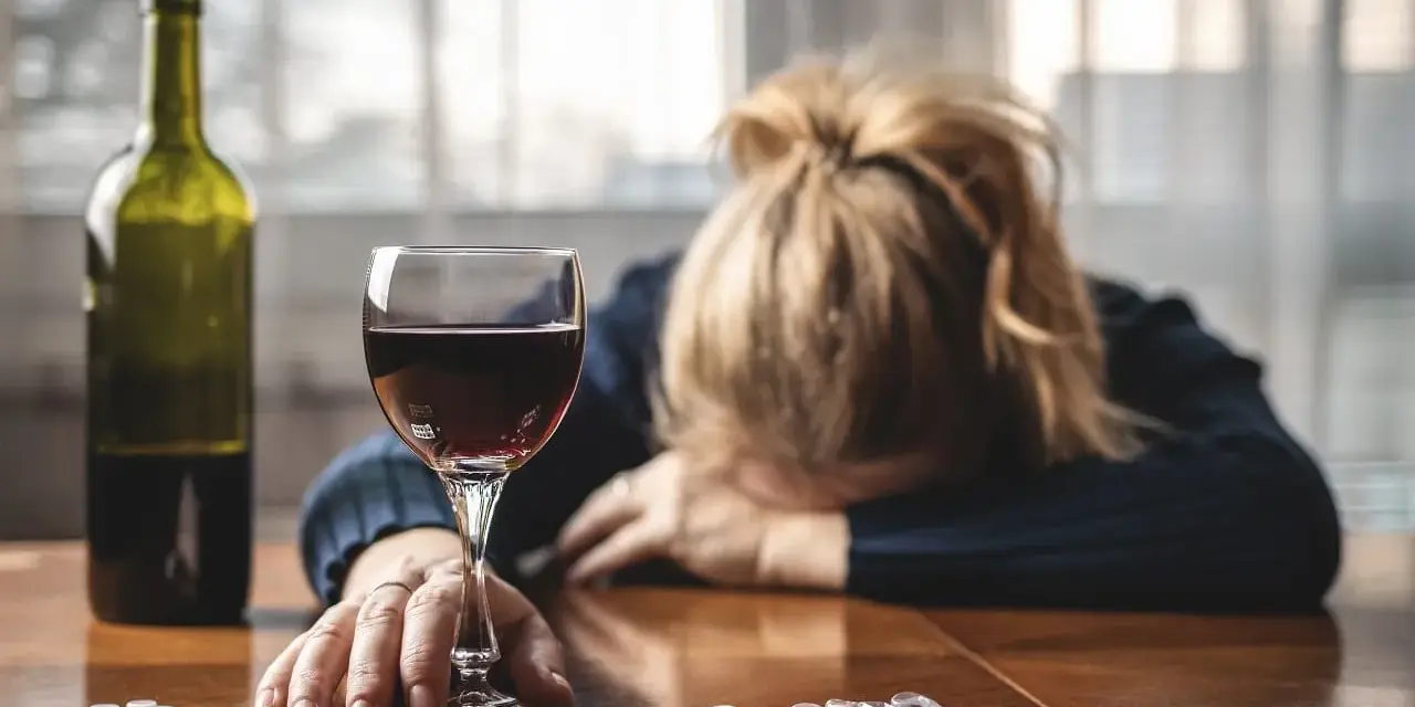 Kobieta z głową opartą na rękach, obok kieliszek czerwonego wina, butelka i opakowania leków, w tym Clexane. Alkohol i leki na stole.