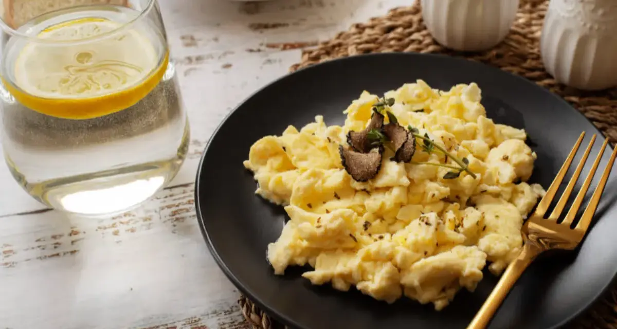 Puszysta jajecznica z truflami na czarnym talerzu, obok szklanka wody z cytryną i kromki chleba. Idealna na śniadanie, znasz jej jajecznica kcal?