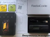Zestaw Radiacode z dozymetrem ZERO, instrukcją i kablem USB. Obok pudełka z urządzeniem znajduje się karta z informacjami o izotopach Co-60 i Sr-90.