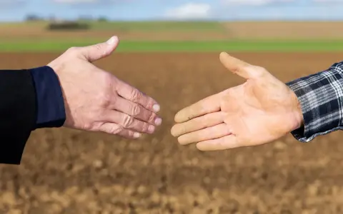 Dwie dłonie, jedna czysta, druga ubrudzona ziemią, zbliżają się do uścisku. To symbol porozumienia między miastem a wsią, biznesu i rolnictwa.