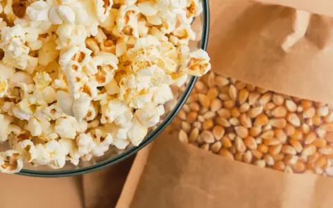 Misternie przygotowany popcorn w szklanej misce obok ziaren kukurydzy w papierowej torbie. Czy popcorn tuczy? Zależy od dodatków!