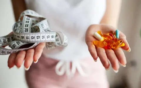 Dwie dłonie: jedna z miarką krawiecką, druga z kolorowymi kapsułkami – symbolizują wybór między dietą a środkami na odchudzanie.