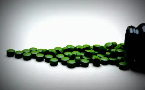 Zielone tabletki chlorelli rozsypane z czarnego pojemnika. Kiedy je brać: chlorella rano czy wieczorem?