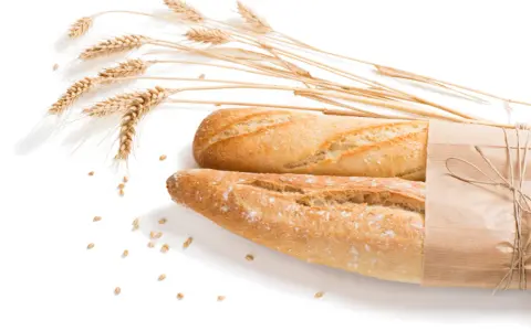 Dwa świeże bagietki, jeden zawinięty w papier, obok kłosów pszenicy i ziaren. Dla osób z alergią na gluten to widok zakazany.