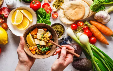 Zdrowa dieta na stawy: miska pełna soczewicy, tofu, warzyw i cytryny. Obok składniki na pyszne danie: pomidory, marchew, awokado, czosnek, cebula, jagody, pesto i chlebki pita.