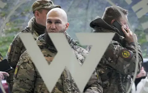 Trzech mężczyzn w mundurach, jeden rozmawia przez telefon, na tle symbolu przypominającego literę "W". Mogą to być członkowie grupy Wagnera.