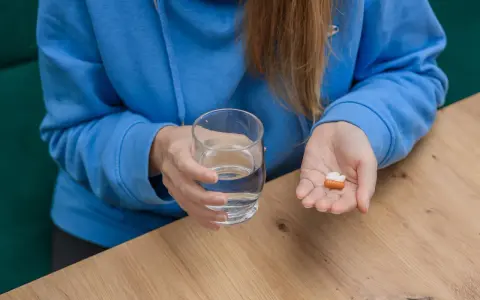 Kobieta w niebieskiej bluzie trzyma szklankę wody i tabletki, w tym cholekalcyferol, na dłoni.