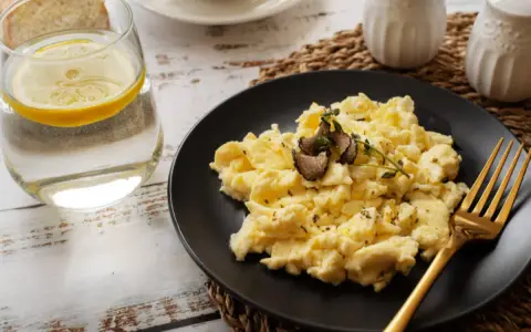 Puszysta jajecznica z truflami na czarnym talerzu, obok szklanka wody z cytryną i kromki chleba. Idealna na śniadanie, znasz jej jajecznica kcal?