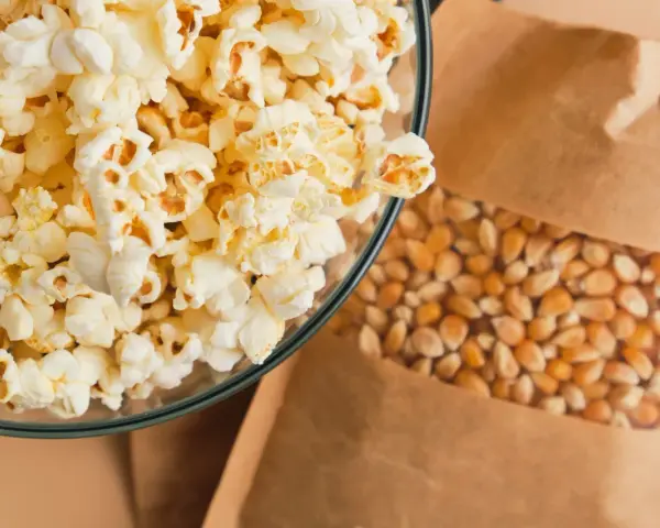 Misternie przygotowany popcorn w szklanej misce obok ziaren kukurydzy w papierowej torbie. Czy popcorn tuczy? Zależy od dodatków!