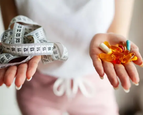 Dwie dłonie: jedna z miarką krawiecką, druga z kolorowymi kapsułkami – symbolizują wybór między dietą a środkami na odchudzanie.