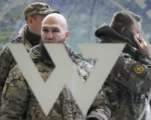 Trzech mężczyzn w mundurach, jeden rozmawia przez telefon, na tle symbolu przypominającego literę "W". Mogą to być członkowie grupy Wagnera.