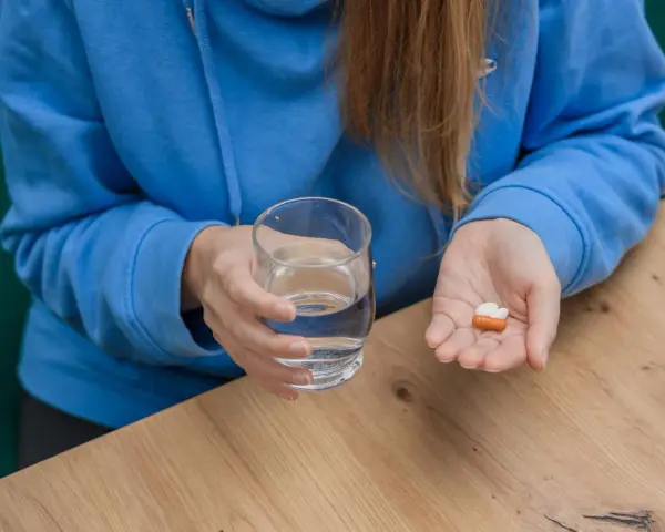 Kobieta w niebieskiej bluzie trzyma szklankę wody i tabletki, w tym cholekalcyferol, na dłoni.