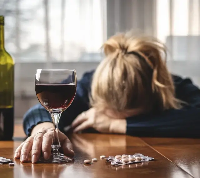 Kobieta z głową opartą na rękach, obok kieliszek czerwonego wina, butelka i opakowania leków, w tym Clexane. Alkohol i leki na stole.