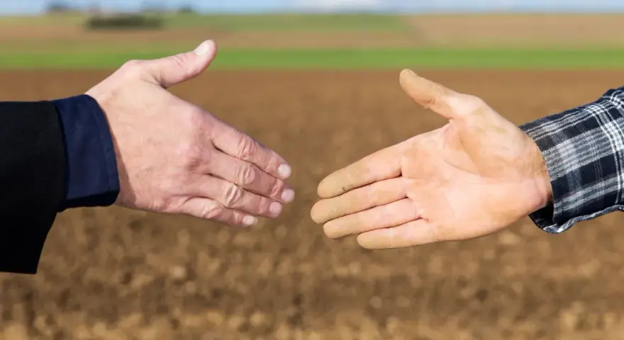 Dwie dłonie, jedna czysta, druga ubrudzona ziemią, zbliżają się do uścisku. To symbol porozumienia między miastem a wsią, biznesu i rolnictwa.