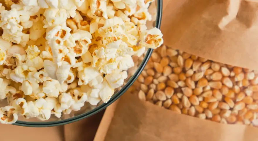 Misternie przygotowany popcorn w szklanej misce obok ziaren kukurydzy w papierowej torbie. Czy popcorn tuczy? Zależy od dodatków!