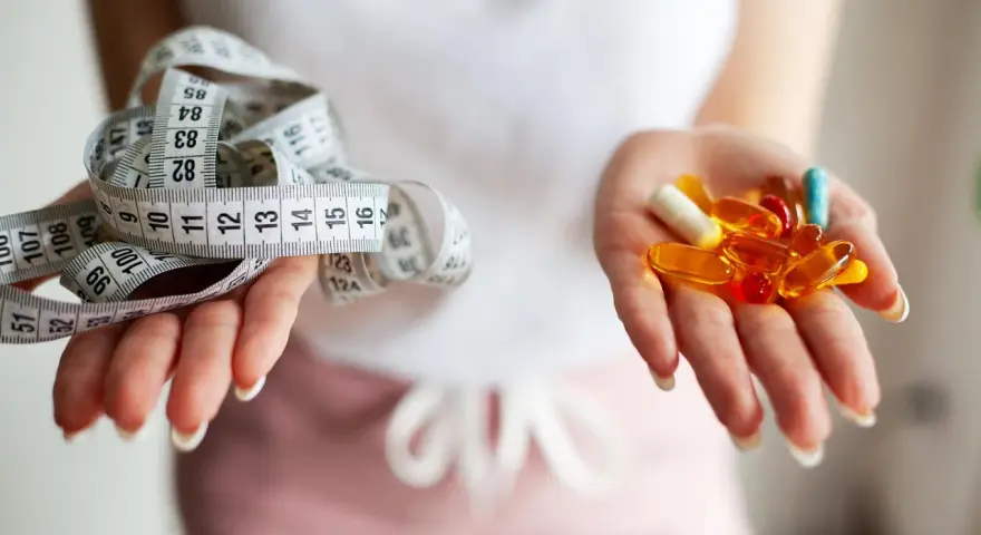 Dwie dłonie: jedna z miarką krawiecką, druga z kolorowymi kapsułkami – symbolizują wybór między dietą a środkami na odchudzanie.