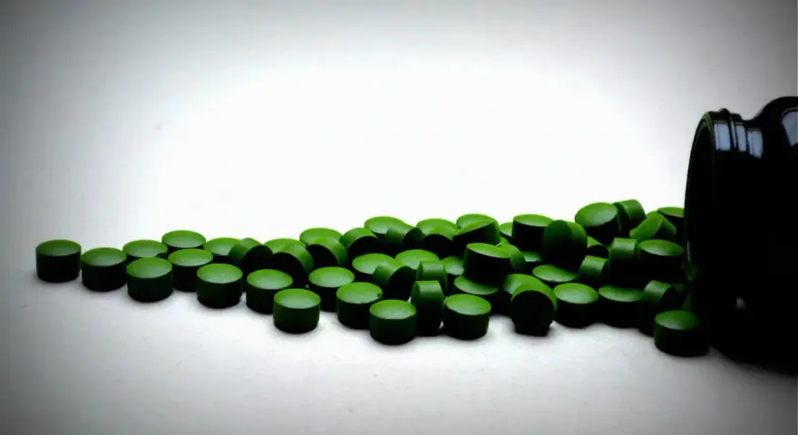 Zielone tabletki chlorelli rozsypane z czarnego pojemnika. Kiedy je brać: chlorella rano czy wieczorem?