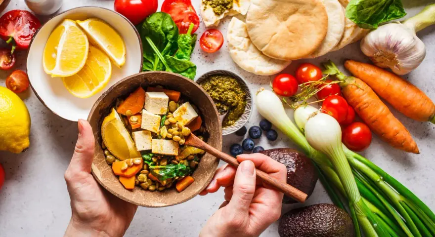 Zdrowa dieta na stawy: miska pełna soczewicy, tofu, warzyw i cytryny. Obok składniki na pyszne danie: pomidory, marchew, awokado, czosnek, cebula, jagody, pesto i chlebki pita.