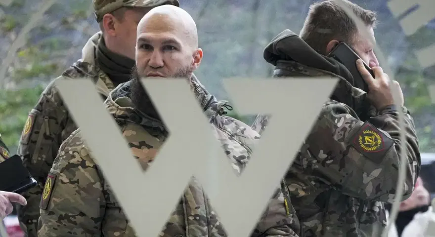 Trzech mężczyzn w mundurach, jeden rozmawia przez telefon, na tle symbolu przypominającego literę "W". Mogą to być członkowie grupy Wagnera.
