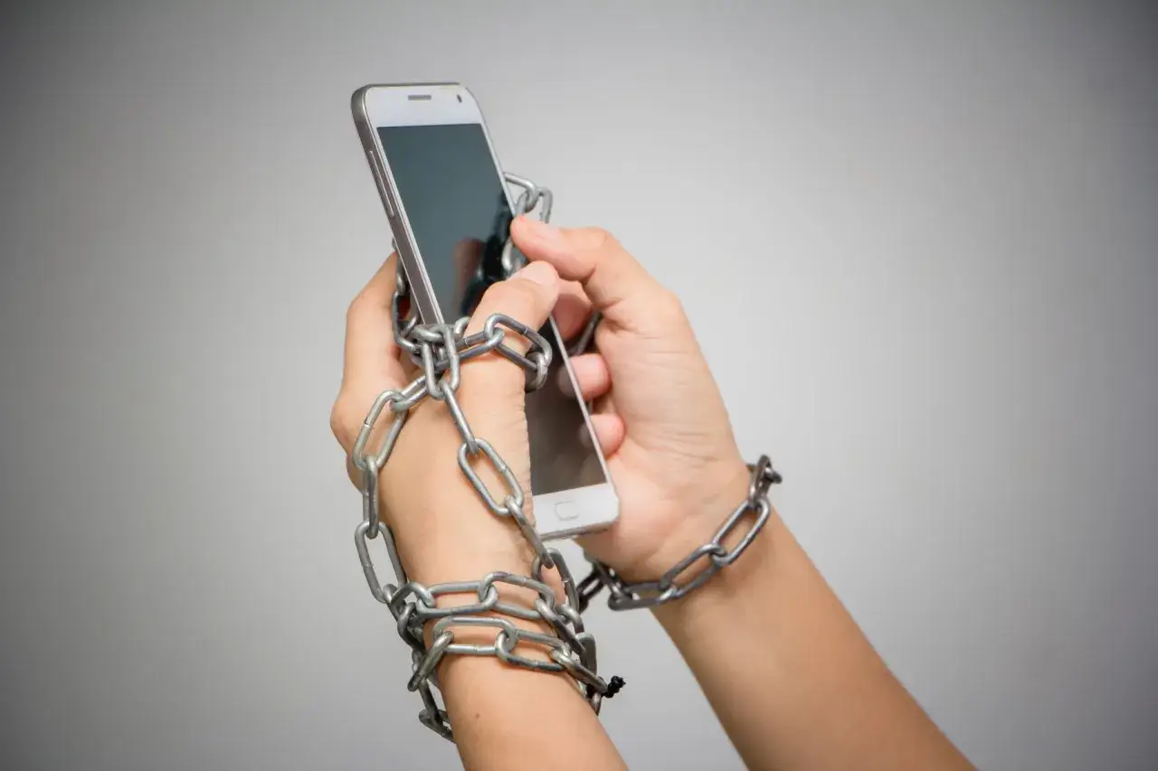 Dłonie spętane łańcuchami, trzymające smartfon. Symbolizuje to nomofobię, lęk przed brakiem telefonu.