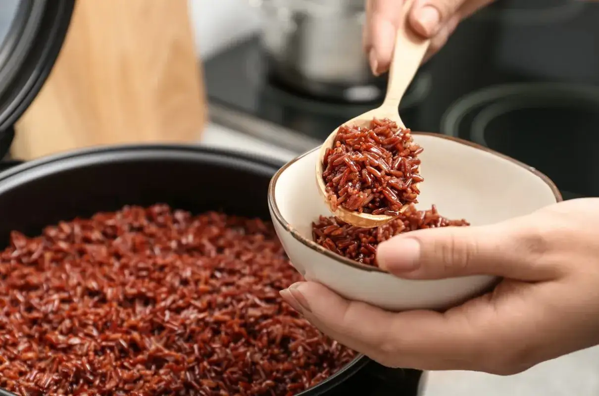 Łyżka nakłada aromatyczny, czerwony ryż do miseczki. W tle garnek z gotującym się ryżem.