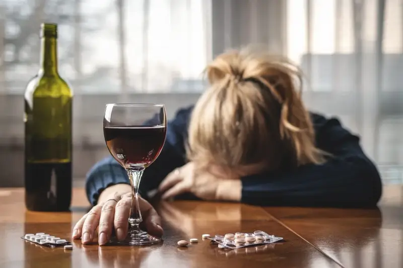 Kobieta z głową opartą na rękach, obok kieliszek czerwonego wina, butelka i opakowania leków, w tym Clexane. Alkohol i leki na stole.